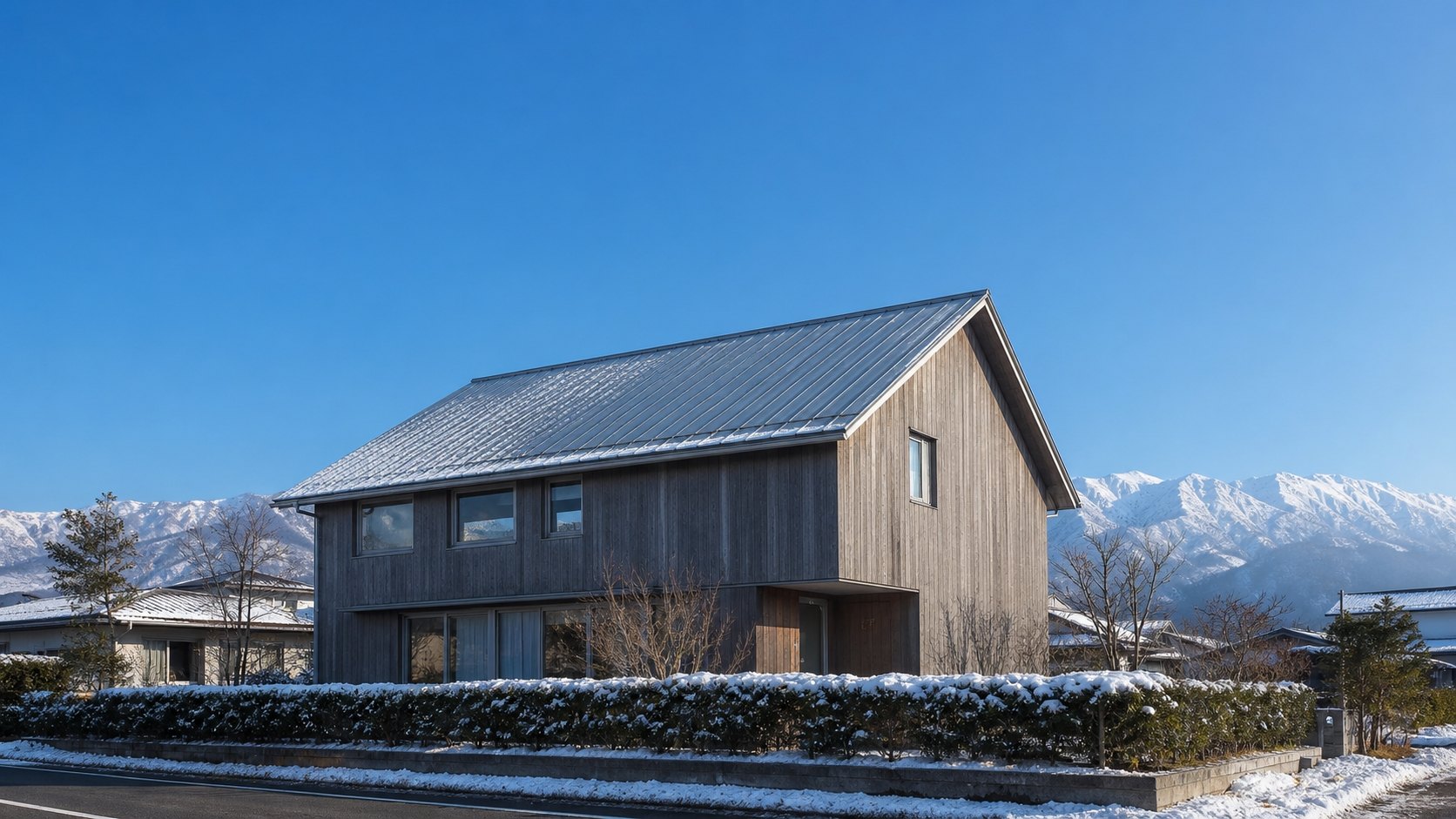 雪をいただいた山並みを背景にした、ガルバリウム鋼板の立平葺きの屋根の住宅